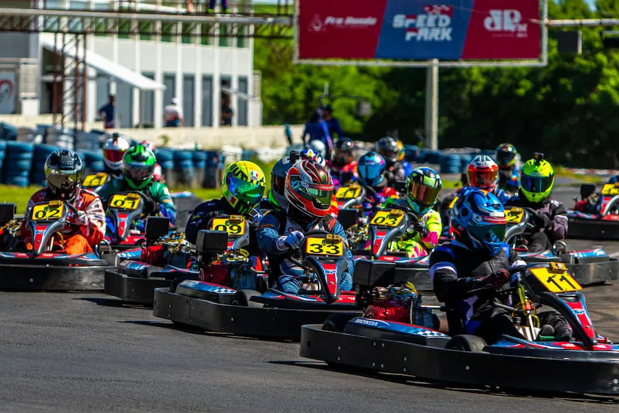 Pilotos de kart em corrida disputando posições lado a lado em pista outdoor, com vários karts alinhados em alta velocidade
