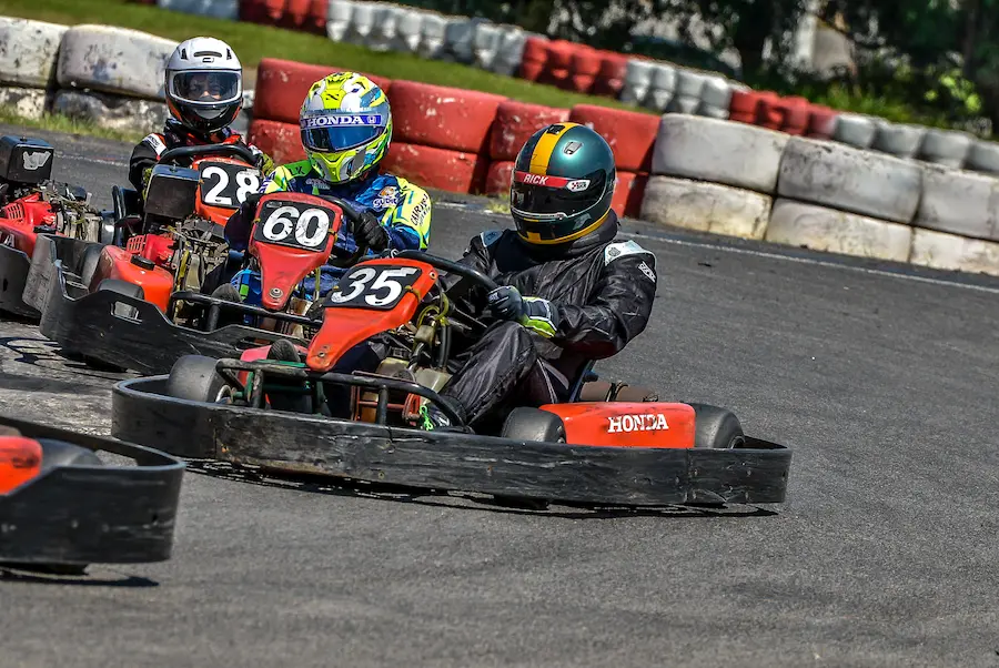 Pilotos de Rental Kart disputando posição em curva durante campeonato, utilizando karts numerados e equipamentos completos de segurança. A imagem destaca a proximidade entre os competidores, o controle em curva e a intensidade típica das corridas de kart em ambiente competitivo.
