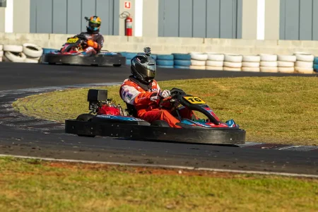 Piloto de kart em pista de kartódromo durante uma corrida, com outro kart ao fundo e barreiras de pneus ao redor do circuito.