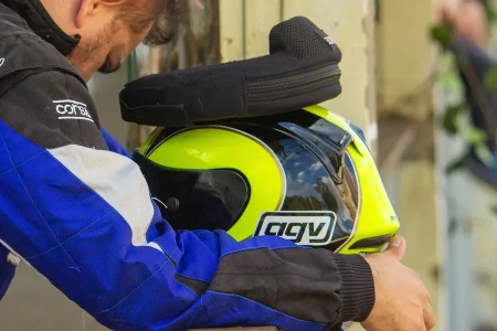 Piloto de kart vestindo macacão ajusta um protetor cervical preto sobre um capacete amarelo e preto antes de entrar na pista. A imagem destaca a preparação e o uso de equipamentos de segurança no kart, reforçando a importância da proteção da região do pescoço durante a pilotagem.