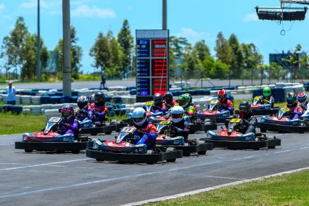 Largada de campeonato de Rental Kart com vários pilotos alinhados na pista, utilizando karts numerados e equipamentos completos de segurança. A imagem mostra o início da corrida em ambiente profissional, com painel de cronometragem ao fundo e disputa intensa entre os participantes.