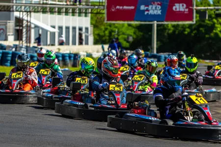 Pilotos de kart em corrida disputando posições lado a lado em pista outdoor, com vários karts alinhados em alta velocidade
